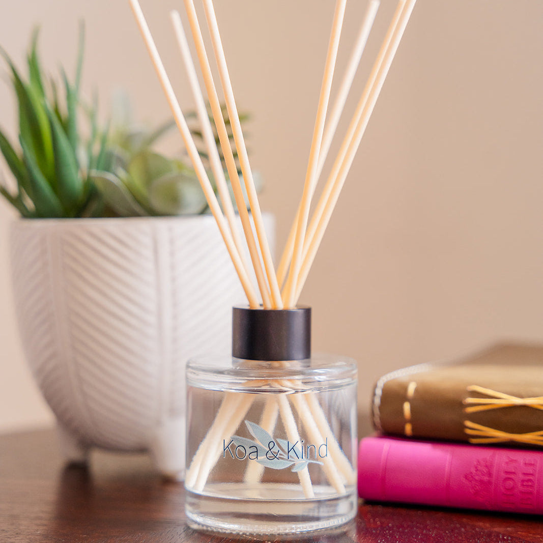Reed diffuser with wooden sticks in a glass container on a wooden surface, with a plant and book in the background.
